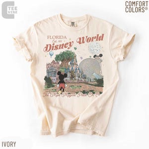 May include: Ivory-colored t-shirt featuring a graphic of Disney World in Florida. The design includes Mickey Mouse, the castle, and the text "Florida Disney World Est. 1971". The shirt is made of Comfort Colors material.