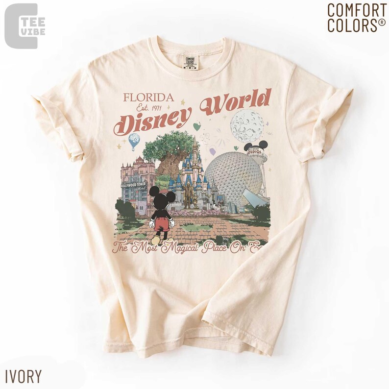May include: Ivory-colored t-shirt featuring a graphic of Disney World in Florida. The design includes Mickey Mouse, the castle, and the text "Florida Disney World Est. 1971". The shirt is made of Comfort Colors material.