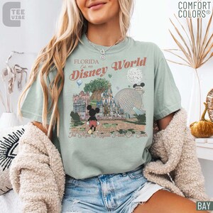 May include: Sage green t-shirt with a graphic of Disney World in Florida. The design includes Mickey Mouse, the Tree of Life, and the Spaceship Earth. Text on the shirt reads "Florida Disney World Est. 1971" and "The Most Magical Place On Earth."