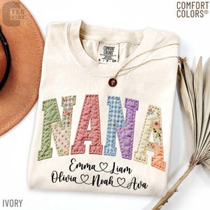May include: Ivory-colored Comfort Colors t-shirt with the word "NANA" in patchwork letters. Below the word are names and hearts. A gold necklace with a pendant rests on the shirt. The shirt is a size small.