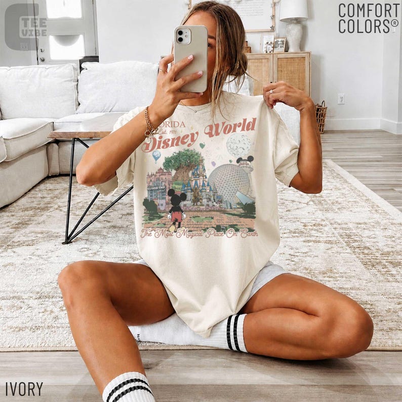 May include: Ivory-colored t-shirt with a graphic of Disney World in Florida. The design includes Mickey Mouse, a castle, and the text "Disney World" and "The Most Magical Place On Earth". The shirt is a Comfort Colors brand.