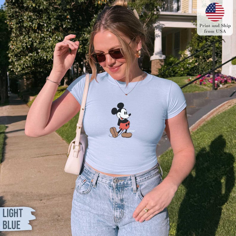 May include: Light blue t-shirt featuring a classic Mickey Mouse graphic. The shirt has short sleeves and a crew neck. The text "LIGHT BLUE" is visible in the bottom left corner. The shirt is paired with light wash denim jeans.