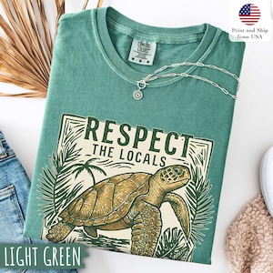 May include: Light green t-shirt with a graphic of a sea turtle and the text "Respect the Locals." The shirt has a crew neck and short sleeves. A silver necklace with a sun pendant is on the shirt. The shirt is labeled "Comfort Colors."