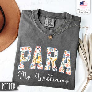Pode incluir: Uma camiseta cinza Comfort Colors com a palavra "PARA" em letras grandes com estampa floral. Abaixo, o nome "Ms. Williams" está escrito em itálico. Um colar de ouro e um chapéu marrom são visíveis.