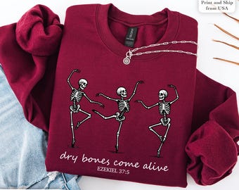 May include: Maroon sweatshirt featuring a graphic of three dancing skeletons. The phrase "dry bones come alive" and Bible verse Ezekiel 37:5 are printed below the skeletons. A silver necklace with a pendant is draped on the sweatshirt.