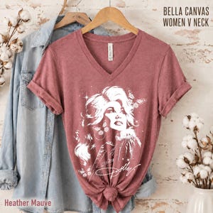 May include: A heather mauve V-neck t-shirt featuring a white graphic illustration of a person with voluminous hair surrounded by butterflies and stars, with the script text Dolly below. The shirt is displayed on a wooden hanger.