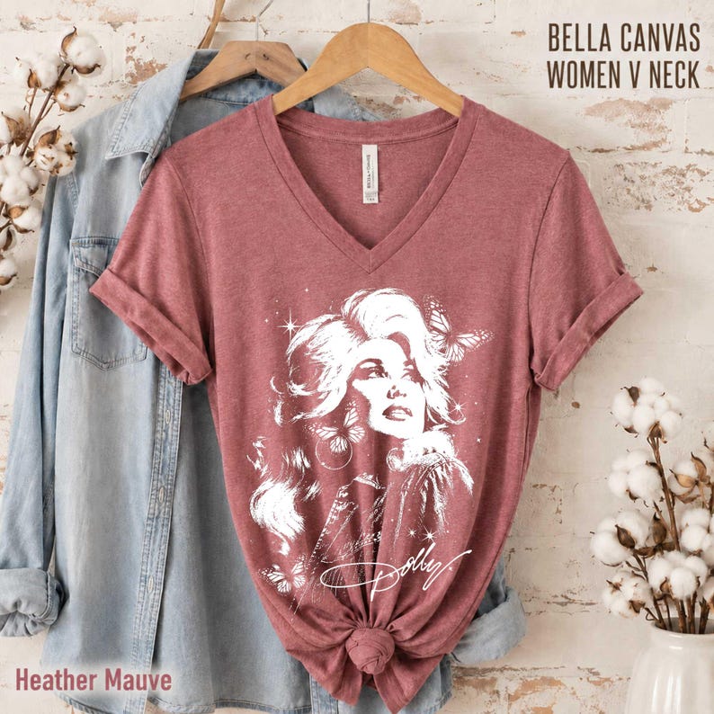 May include: A heather mauve V-neck t-shirt featuring a white graphic illustration of a person with voluminous hair surrounded by butterflies and stars, with the script text Dolly below. The shirt is displayed on a wooden hanger.