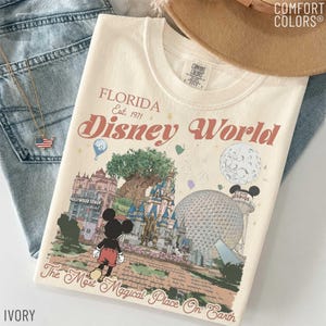 May include: Ivory-colored t-shirt with a graphic of Disney World in Florida. The design includes the text "Florida Disney World Est. 1971" and illustrations of iconic park features, including Mickey Mouse. The shirt is paired with blue jeans and a gold necklace.