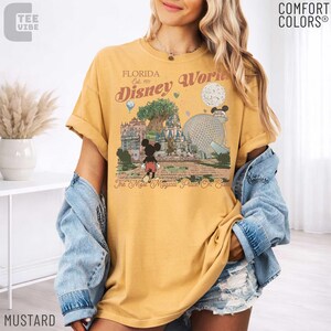 May include: Mustard-colored t-shirt featuring a vintage-style Disney World graphic. The design includes Mickey Mouse, a castle, and the text "Florida Disney World Est. 1971". The shirt is a Comfort Colors brand.