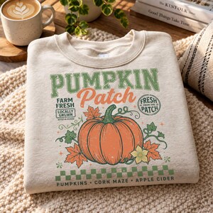 May include: A beige crewneck sweatshirt featuring a graphic print of a large orange pumpkin surrounded by autumn leaves and vines. The text reads PUMPKIN Patch, FARM FRESH LOCALLY GROWN, FRESH FROM THE PATCH, PUMPKINS, CORN MAZE, and APPLE CIDER.