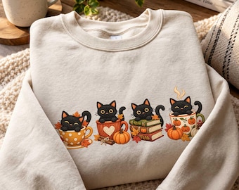 May include: A beige crewneck sweatshirt featuring a graphic print of four black cats. One cat sits in a yellow polka-dot teacup, another in an orange heart-patterned mug, one rests on a stack of books, and the last sits in a pumpkin-patterned mug.