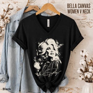 May include: A black V-neck t-shirt featuring a white graphic illustration of a woman with voluminous hair, surrounded by butterflies and stars, with the script text Dolly. The shirt is displayed on a wooden hanger alongside a light blue denim shirt.