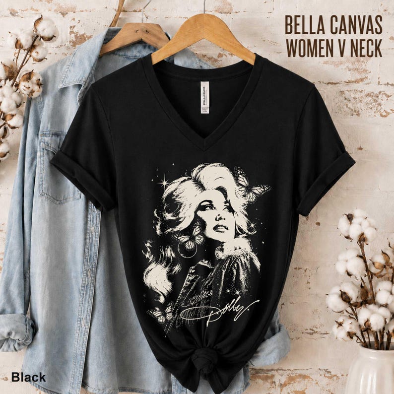 May include: A black V-neck t-shirt featuring a white graphic illustration of a woman with voluminous hair, surrounded by butterflies and stars, with the script text Dolly. The shirt is displayed on a wooden hanger alongside a light blue denim shirt.