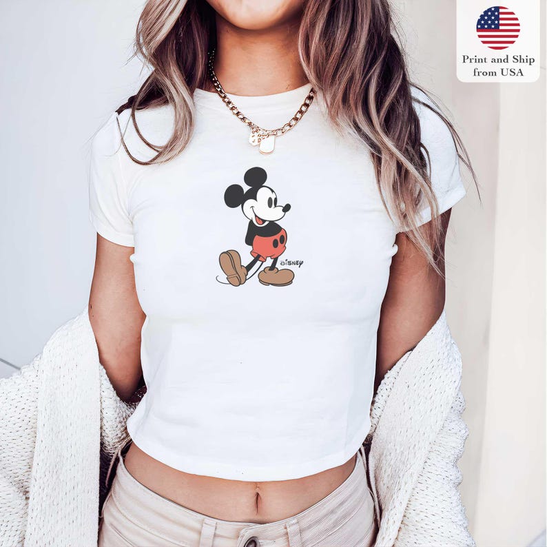 May include: White crop top featuring a classic Mickey Mouse graphic. The cartoon character is depicted in black, red, and brown, with the Disney logo below. The shirt is short-sleeved and has a round neckline. The image also includes the text "Print and Ship from USA".