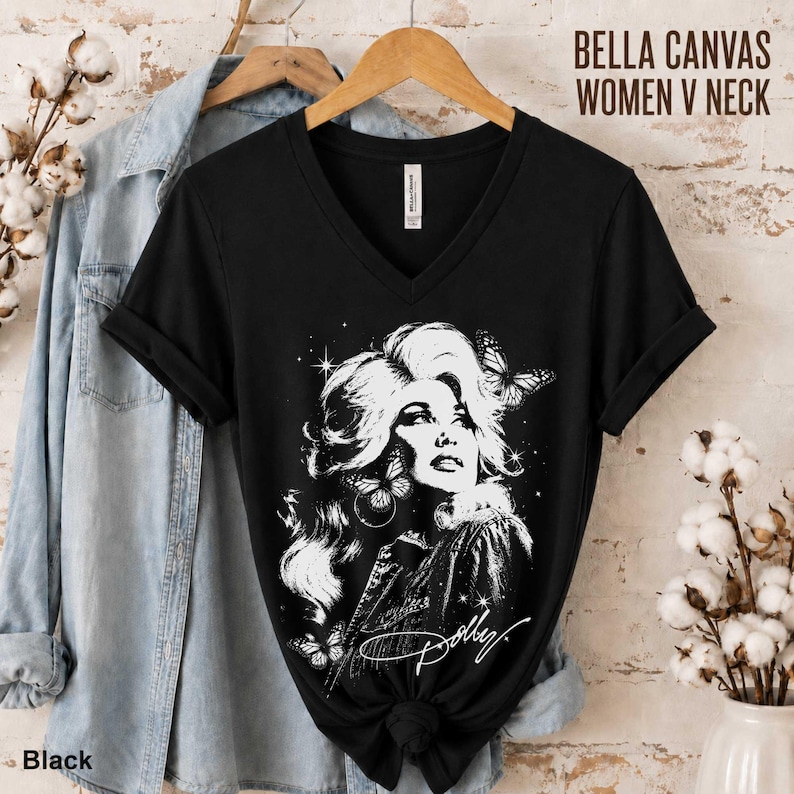 May include: A black V-neck t-shirt featuring a white graphic illustration of a person with voluminous hair, butterflies, and stars. The text "Dolly" is written in a script font at the bottom of the graphic. The shirt is displayed on a wooden hanger.