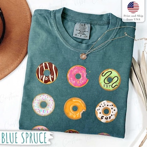 May include: Blue spruce Comfort Colors t-shirt with a donut graphic. The donuts are brown, pink, green, white, and orange, each with the letters "FDT". The shirt has a crew neck and short sleeves. A gold necklace is visible.