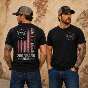 May include: Two black t-shirts with the American flag design. One shirt features the flag on the back with "1776", "We the People", and "250 Years 2026". The other shirt has "1776" in a circle of stars on the front. Both shirts are worn with a baseball cap.