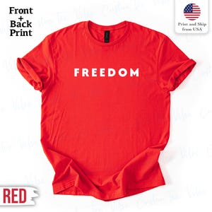 Op de afbeelding: Een felrood t-shirt met het woord "FREEDOM" in witte hoofdletters. Het shirt heeft een ronde hals en korte mouwen. De afbeelding bevat ook een kleine afbeelding van de Amerikaanse vlag.