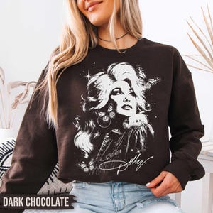 May include: A dark chocolate brown crewneck sweatshirt featuring a white graphic print of a woman with voluminous hair surrounded by butterflies and stars. The script text Dolly is printed in white at the bottom of the graphic design.
