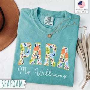 Pode incluir: Camiseta verde-água com a palavra "PARA" em estampa floral e o nome "Ms. Williams" em escrita branca. Um colar de ouro repousa sobre a camisa. A camisa é da Comfort Colors e é tamanho P.