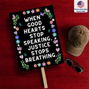 Puede incluir: Un letrero negro con el texto "WHEN GOOD HEARTS STOP SPEAKING, JUSTICE STOPS BREATHING." est&aacute; decorado con dise&ntilde;os florales y de coraz&oacute;n. Una gorra marr&oacute;n y gafas de sol descansan sobre una superficie roja.