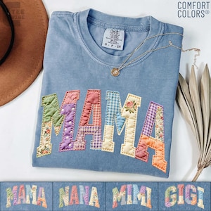 May include: A blue Comfort Colors t-shirt with the word "MAMA" appliqued in colorful patchwork fabric. The letters are outlined in yellow stitching. A gold necklace with a pendant rests on the shirt.