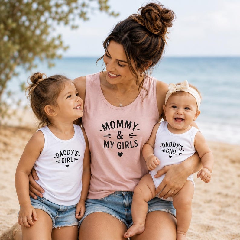 以下が含まれることがあります： MOMMY & MY GIRLSと黒い文字で書かれた薄いピンクのタンクトップと、DADDY'S GIRLと黒い文字で書かれた2枚の白いタンクトップ。衣類にはテキストの下にハートのグラフィックが施されています。白いタンクトップは袖なしのデザインです。
