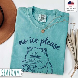 Puede incluir: Camiseta verde mar con la frase "no ice please" y un dibujo de un gato sosteniendo un vaso de hielo. La camiseta tiene cuello redondo y mangas cortas. Un collar dorado con un colgante est&aacute; sobre la camiseta.