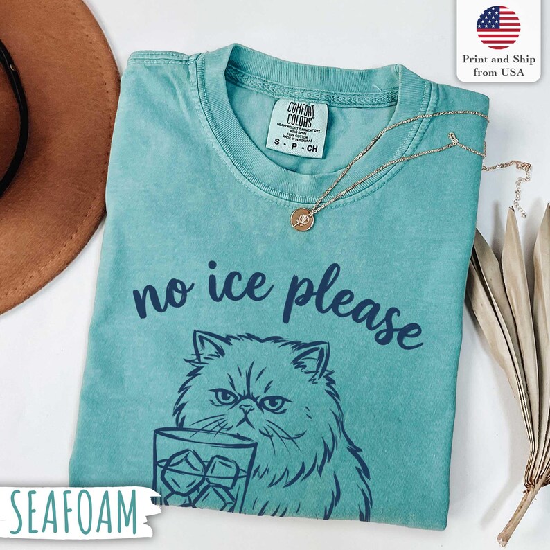 Puede incluir: Camiseta verde mar con la frase "no ice please" y un dibujo de un gato sosteniendo un vaso de hielo. La camiseta tiene cuello redondo y mangas cortas. Un collar dorado con un colgante est&aacute; sobre la camiseta.