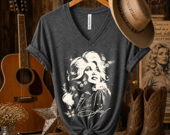 May include: A dark gray V-neck t-shirt featuring a white graphic illustration of a woman with voluminous hair, butterflies, and the script text Dolly. The shirt is displayed on a wooden hanger with a tan cowboy hat resting on the hanger hook.