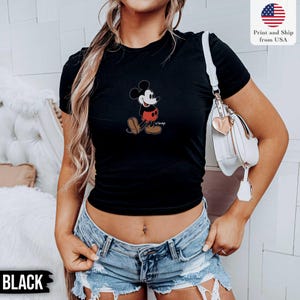 May include: Black t-shirt featuring a classic Mickey Mouse graphic. The cartoon character is depicted in his iconic pose, wearing red shorts, brown shoes, and white gloves. The word "Disney" is printed below the character. The text "Print and Ship from USA" is also visible.
