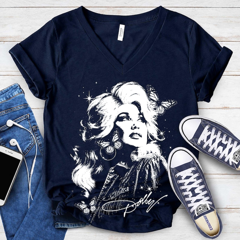 May include: A navy blue V-neck t-shirt featuring a white graphic print of a woman with voluminous hair, butterflies, and stars. The shirt includes the signature text Dolly in white script at the bottom, paired with blue jeans and sneakers.