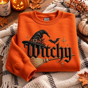 Op de afbeelding: Een oranje sweater met ronde hals en een grafische print van een zwarte heksenhoed, een vliegende vleermuis en een bezemsteel. Het woord "Witchy" staat in het midden gedrukt in een vet, zwart gotisch lettertype, geaccentueerd met gouden sterren.