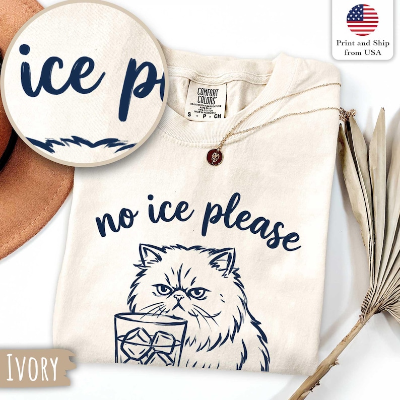 Puede incluir: Camiseta de color marfil con la frase "no ice please" y un gr&aacute;fico de un gato gru&ntilde;&oacute;n sosteniendo un vaso de hielo. La camiseta tambi&eacute;n tiene las palabras "ice p". Se ve un collar dorado con un colgante.