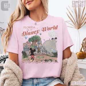 May include: Pink t-shirt with a graphic of Disney World in Florida. The design includes the text "Florida Disney World Est. 1971" and a cartoon Mickey Mouse. The shirt is labeled "Comfort Colors" and "Blossom".