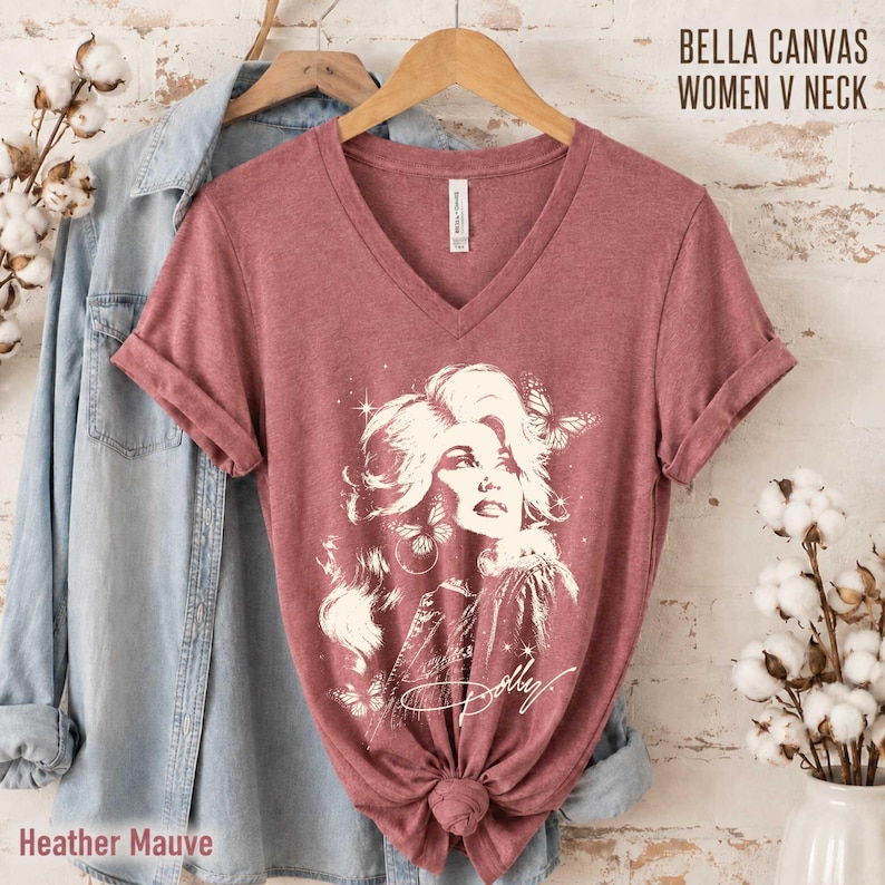 May include: A heather mauve V-neck t-shirt featuring a white graphic illustration of a person with voluminous hair surrounded by butterflies and stars. The shirt includes the text BELLA CANVAS WOMEN V NECK and Heather Mauve.