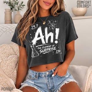 Może przedstawiać: Ciemnoszary t-shirt z krótkim rękawem z białym napisem graficznym Ah! The Element of Surprise, symbolem atomu oraz ilustracją kolby laboratoryjnej. Koszulka zawiera również napis Comfort Colors.