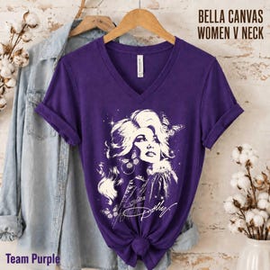 May include: A purple V-neck t-shirt featuring a white graphic illustration of a person with voluminous hair, butterflies, and stars. The shirt includes the text BELLA CANVAS WOMEN V NECK and the script signature Dolly. A light blue denim shirt is layered underneath.