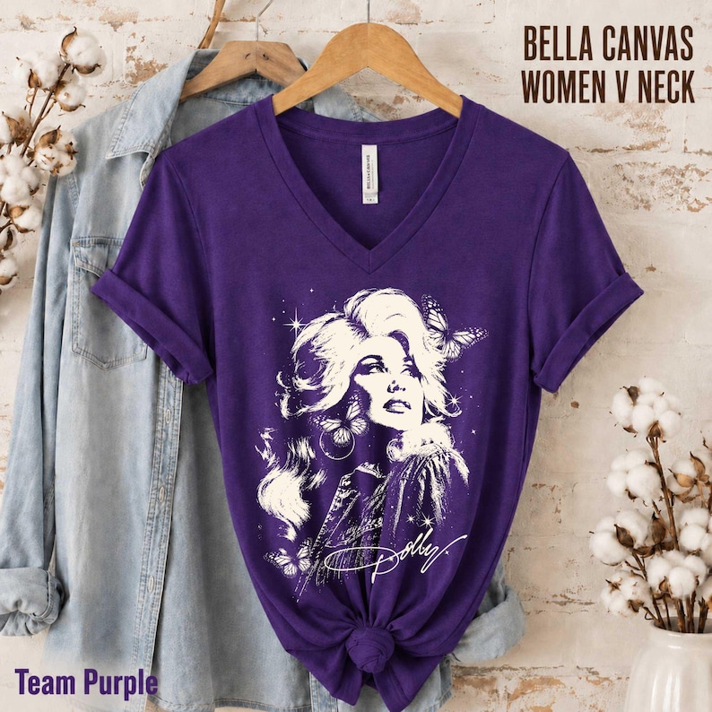 May include: A purple V-neck t-shirt featuring a white graphic illustration of a person with voluminous hair, butterflies, and stars. The shirt includes the text BELLA CANVAS WOMEN V NECK and the script signature Dolly. A light blue denim shirt is layered underneath.