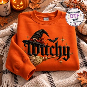 May include: An orange crewneck sweatshirt featuring a black witch hat, a broomstick, a flying bat, and the word Witchy in a gothic font. The design includes small star accents and a circular sticker stating High Quality DTF Print Not Embroidery.
