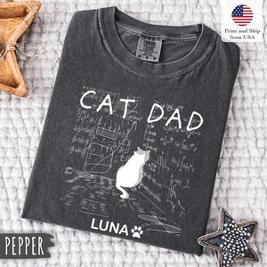 May include: Dark gray t-shirt with the words "CAT DAD" in white, along with a white cat illustration and the name "LUNA". The shirt also features a paw print and mathematical equations.