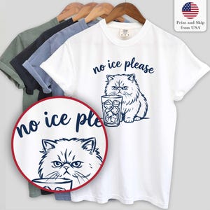 Puede incluir: Camiseta blanca con una ilustraci&oacute;n azul marino de un gato persa y un vaso de cubitos de hielo. El texto "no ice please" est&aacute; encima del gato. Se ven otras camisetas de diferentes colores.