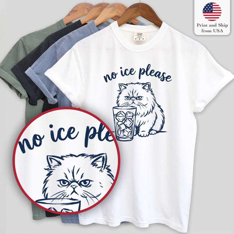 Puede incluir: Camiseta blanca con una ilustraci&oacute;n azul marino de un gato persa y un vaso de cubitos de hielo. El texto "no ice please" est&aacute; encima del gato. Se ven otras camisetas de diferentes colores.