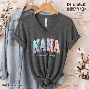 May include: A dark heather gray V-neck t-shirt featuring the word NANA in a colorful, multi-patterned quilt-style font. Below the word, the names Olivia and Lucas are printed in a white script font with a small heart symbol in between.