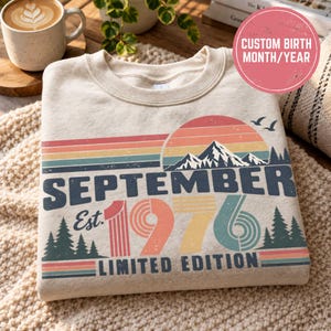 Può includere: Una felpa girocollo beige con una stampa grafica raffigurante un tramonto, una catena montuosa e alberi di pino. Il testo recita SEPTEMBER Est. 1976 LIMITED EDITION in blu scuro e colori tenui, con due uccelli che volano vicino al sole.