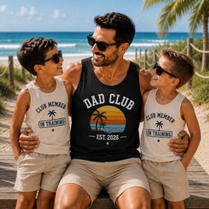 以下が含まれることがあります： 夕日のグラフィックと「DAD CLUB EST. 2026」の文字が入った黒のタンクトップと、「CLUB MEMBER IN TRAINING」の文字とヤシの木のグラフィックが入ったベージュのタンクトップ2枚。3着ともラウンドネックで袖なしのデザインです。