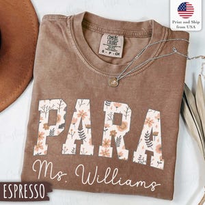 Pode incluir: Camiseta marrom com a palavra "PARA" em letras grandes com estampa floral. O nome "Ms. Williams" está escrito em itálico abaixo. Um colar de prata com um pingente repousa sobre a camiseta. A camiseta tem uma etiqueta "Comfort Colors".