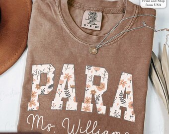 May include: Espresso-colored t-shirt with the word "PARA" in large, floral-patterned letters. Below, the name "Ms. Williams" is written in cursive. A silver necklace with a pendant rests on the shirt. The shirt has a "Comfort Colors" tag.