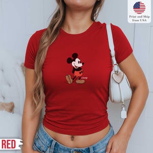 May include: A red t-shirt featuring a classic Mickey Mouse graphic. The cartoon character is depicted in his iconic pose, with black ears, a white face, red shorts, and brown shoes. The shirt also has the text "Disney" below the character. The text "Print and Ship from USA" is also visible.