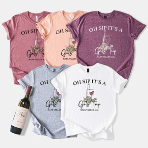 May include: Five short-sleeved t-shirts in shades of pink, peach, gray, and white. Each shirt features the text "OH SIP IT'S A Girls Trip NAPA VALLEY 2025" with a wine bottle and glass graphic. A wine bottle is visible.
